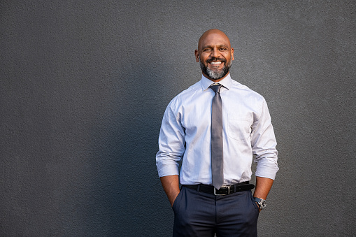 Sponsored image Portrait of successful black businessman standing against grey wall. Handsome senior entrepreneur wearing blue tie and formal clothing smiling and looking at camera. Mature happy man with hands in pocket isolated on grey background with copy space.